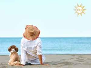 Back of young woman wearing a hat and long sleeved white shirt for sun protection and her small brown and white dog sitting on a beach in cloudless sunny sky facing the water.