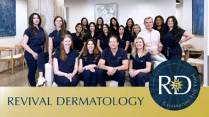 A group of people in navy blue uniforms and business casual attire pose indoors with a Revival Dermatology Dallas anniversary logo in the foreground. | Revival Dermatology, Dallas TX